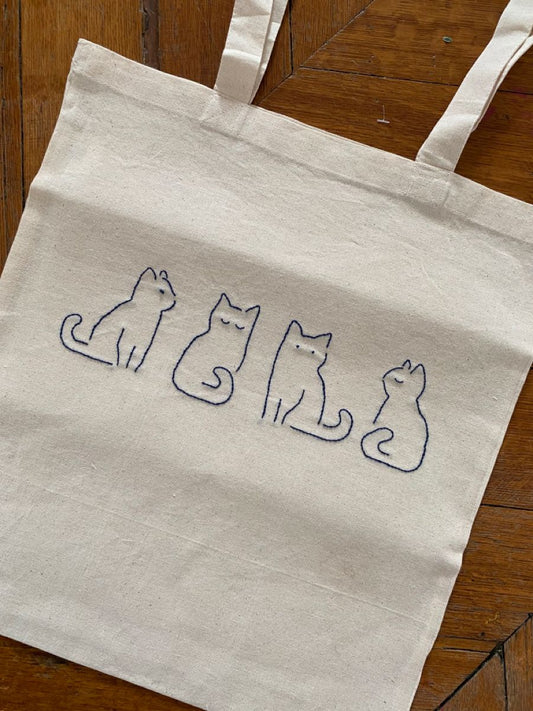 Beige tote bag with embroidered cat designs on a wooden surface